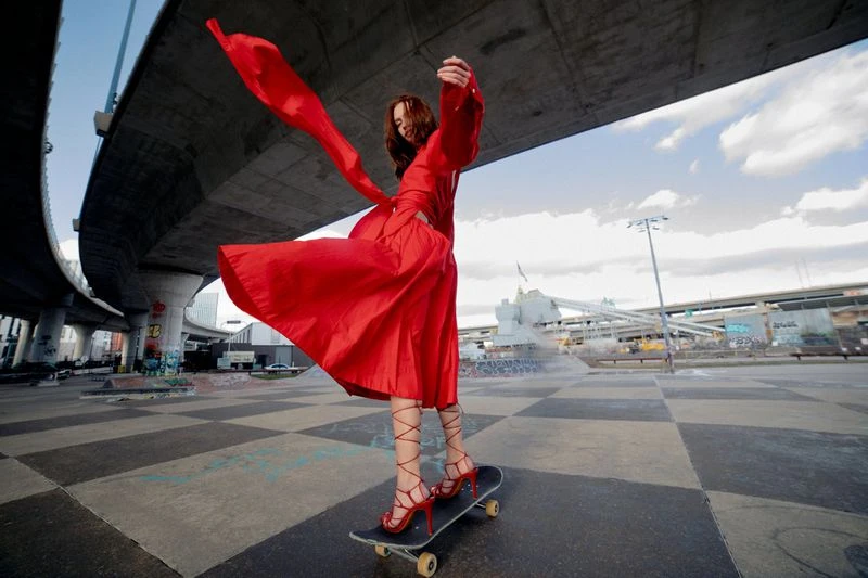 A woman in a red dress is skateboarding at an outdoor skatepark in front of a concrete bridge, wearing high heels and posing. 