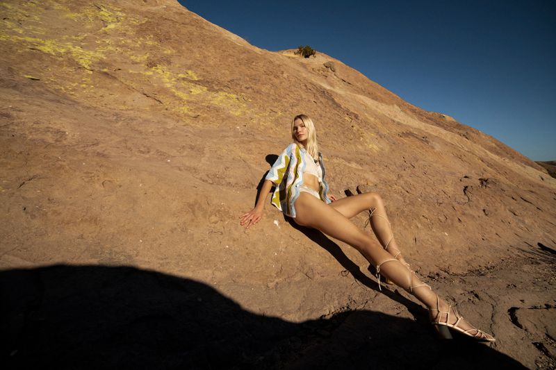 Woman in shirt and lace-up sandals lounges on warm-toned rock formation under clear blue sky on a sunny day in an outdoor setting.