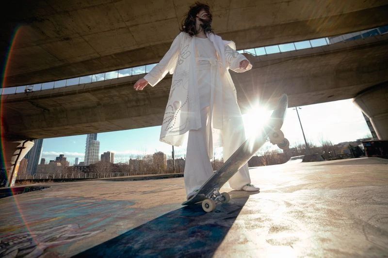 A young adult woman in an all-white outfit stands on a skateboard, outdoors in an urban environment, under a bridge on a sunny day.