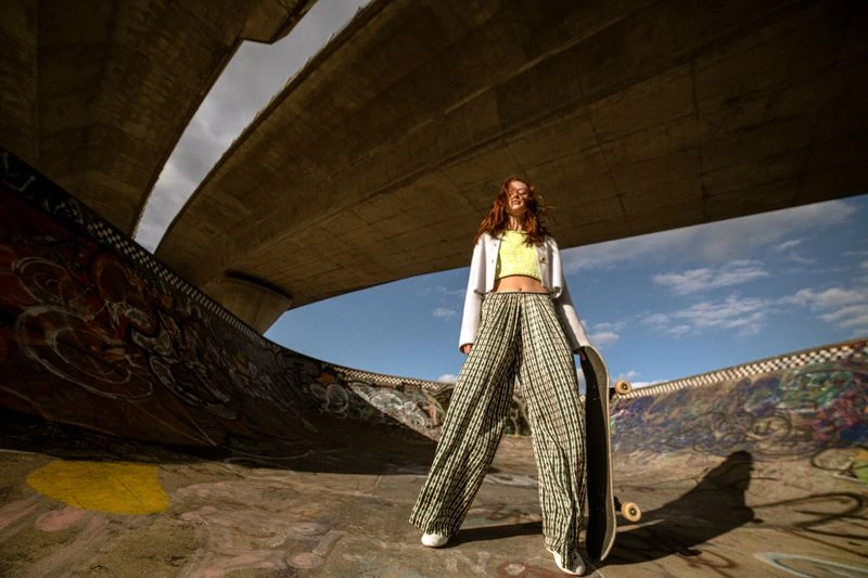 Stylish redhead woman poses in a skatepark with a skateboard. She wears loose pants, a cropped top, and a jacket against a cloudy sky.