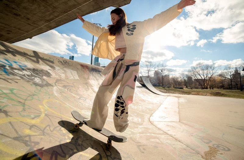 Young woman skateboarding in skatepark wearing patterned jacket and pants, blue sky and city buildings visible in the distance.