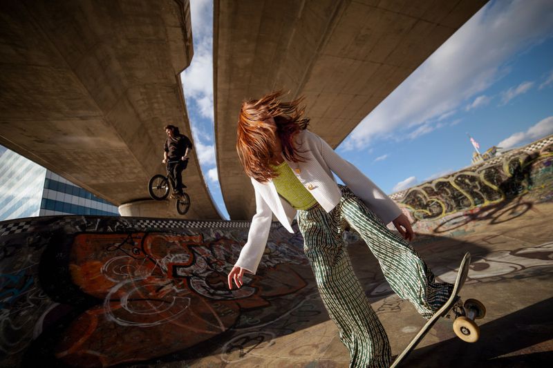 Young woman skateboarding in an urban skatepark with graffiti, under a bridge with a cyclist on a BMX bike. Sunny day, dynamic composition.