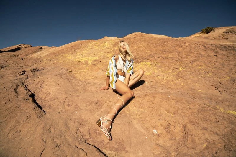 A young blonde woman is sitting on a rocky landscape wearing a matching shirt and shorts with strappy heeled sandals against blue sky.