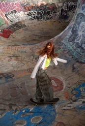 Red-haired female skateboarder riding down a graffiti-covered concrete ramp wearing patterned trousers and white jacket