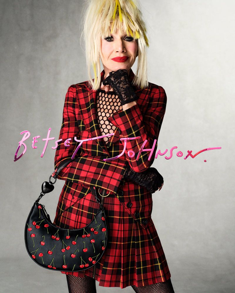 A mature woman in a red plaid blazer dress and black fishnet stockings poses with a cherry-patterned black purse on a grey background.