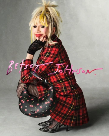 Woman in a red plaid coat posing with a black cherry-print handbag, seated indoors against a grey background, looking at the camera.