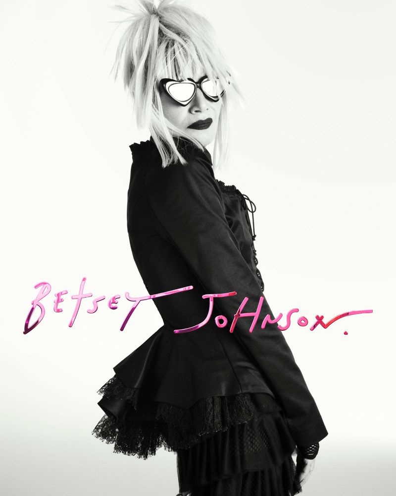 Fashion shot of a woman wearing heart-shaped glasses, dark makeup, and a black dress, posing in a bright photo studio with signature text overlay.