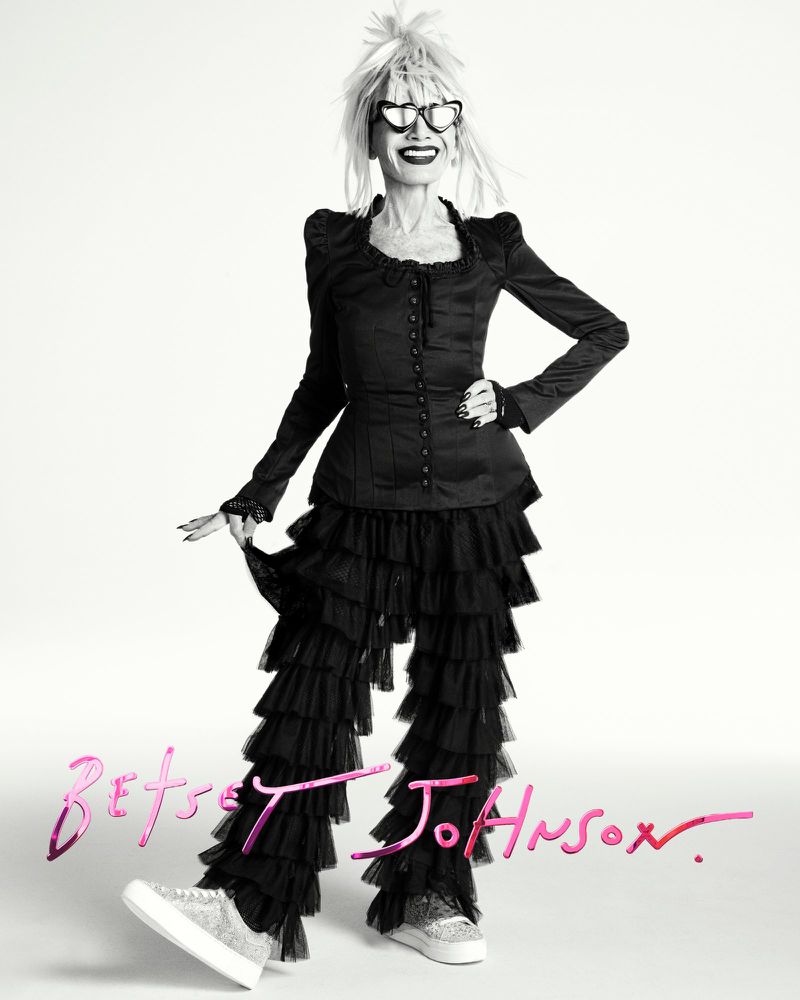 Mature woman with blonde hair and heart sunglasses wears a black ruffle pantsuit and glitter sneakers in a studio shot.