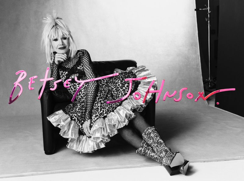 A seated woman poses in a leopard-print dress and fishnet stockings against a grayscale studio backdrop, accompanied by the pink script, Betsey Johnson.