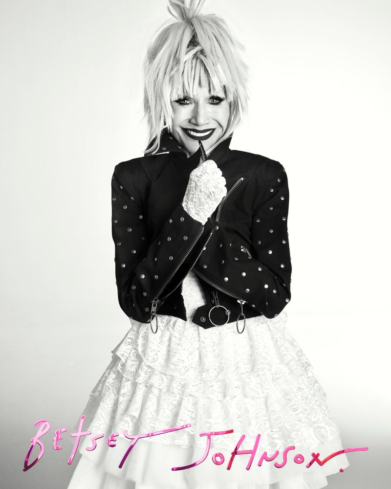A person in a black and white portrait wearing a black studded jacket and a white layered ruffle skirt with blond spiky hair.