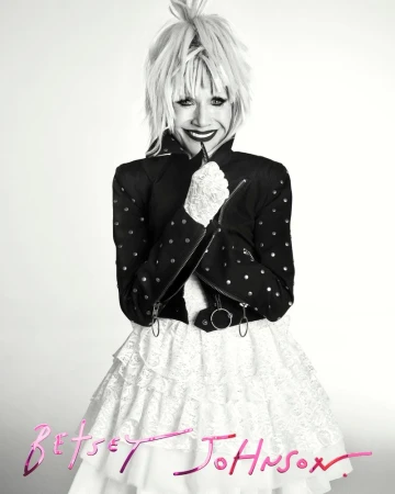 A person in a black and white portrait wearing a black studded jacket and a white layered ruffle skirt with blond spiky hair.
