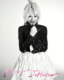 Betsey Johnson smiling in black studded jacket and white lace dress against a white seamless background for Spring Campaign