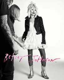 Betsey Johnson standing in white studio wearing a white dress, black jacket, fishnet tights, and silver cowboy boots.
