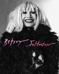 Portrait of Betsey Johnson smiling with blonde fringe wig in black textured coat against gradient background