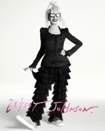 Betsey Johnson wearing a black ruffled outfit and cat-eye glasses against a white backdrop