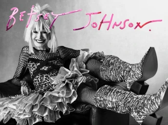 Monochrome portrait of Betsey Johnson lounging on a leather chair wearing a leopard print dress and tall patterned boots