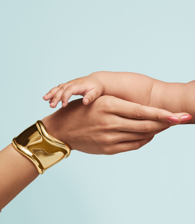 Close-up studio shot of an adult's hand wearing a gold bracelet holding a baby's hand, against a pale blue background.