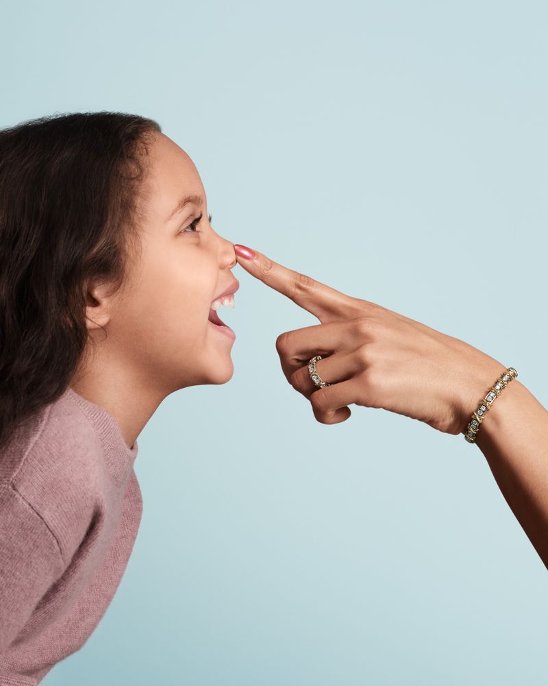 A smiling child has her nose touched by a finger wearing rings against a light blue background in a well-lit, eye-level studio shot.