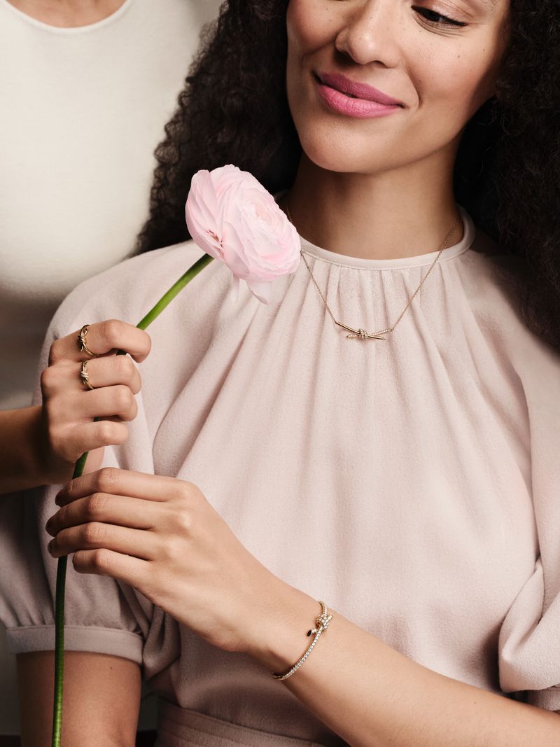 Close-up shot of a smiling woman with dark hair wearing jewelry and holding a pink flower, soft studio lighting, and a second person.