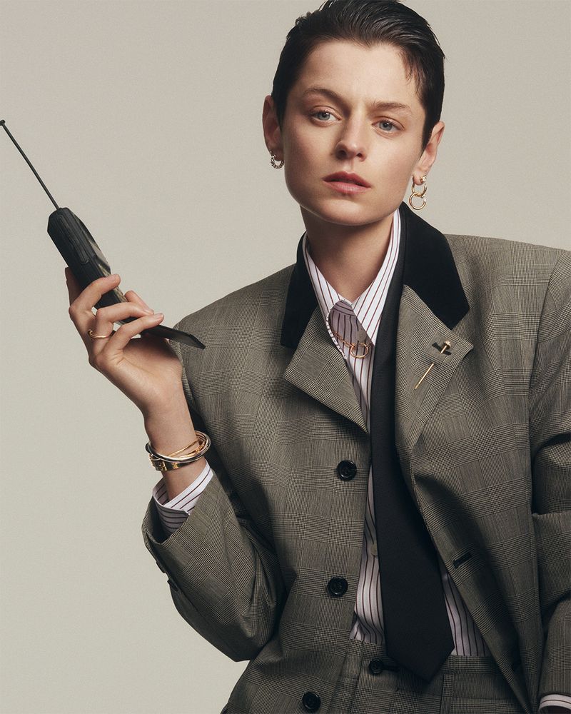 Emma Corrin poses indoors holding a vintage telephone, wearing a gray suit jacket, white striped shirt, and black tie in a studio shot.