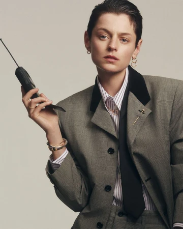 Emma Corrin poses indoors holding a vintage telephone, wearing a gray suit jacket, white striped shirt, and black tie in a studio shot.