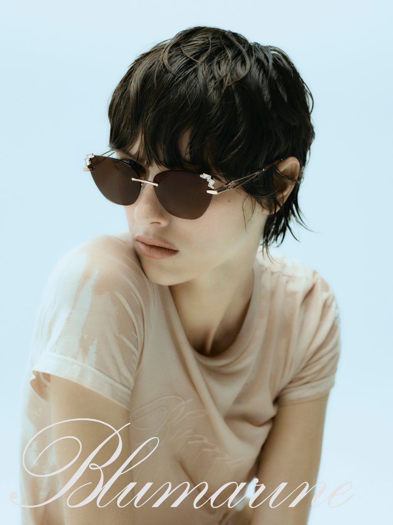 Close-up studio portrait of an adult female model wearing sunglasses and a wet t-shirt against a light blue background. 