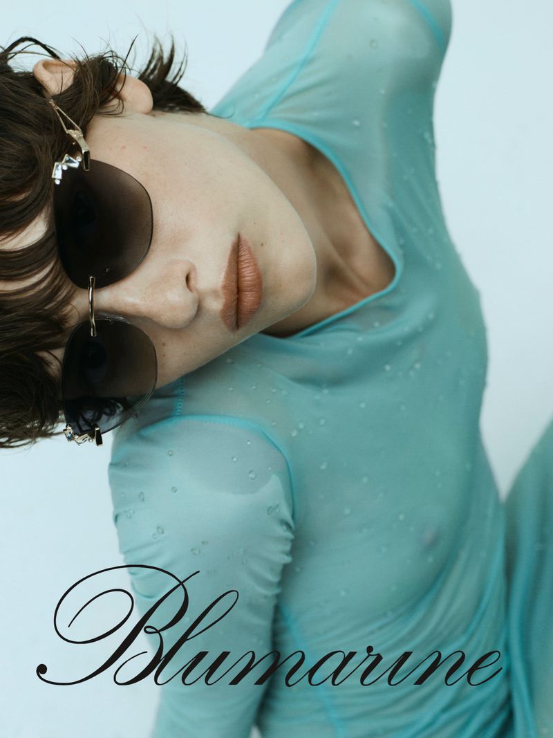 Close-up fashion shot of a woman with brunette hair wearing sunglasses and a light blue transparent long-sleeve top against a white backdrop.