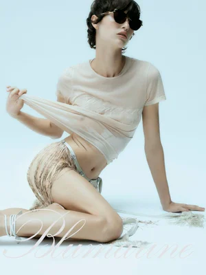 Model wearing dark sunglasses and beige top lifting hem against light background