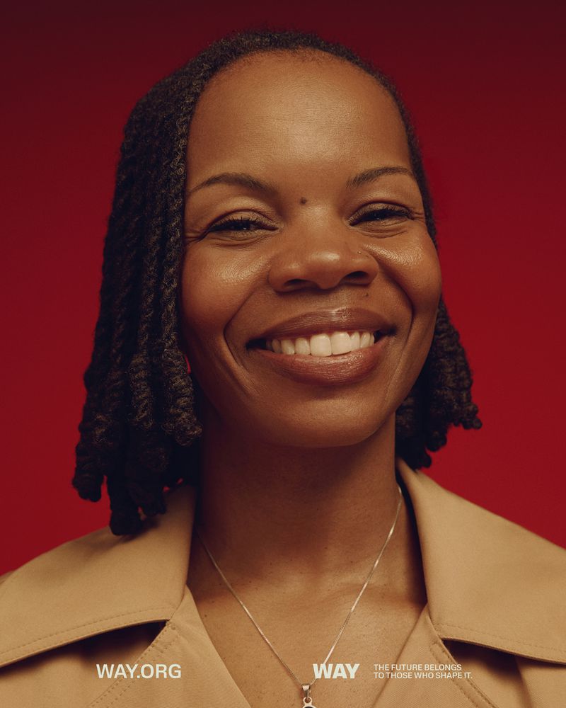 Close-up portrait of a smiling African-American woman with braided hair wearing a tan coat in front of a red background.