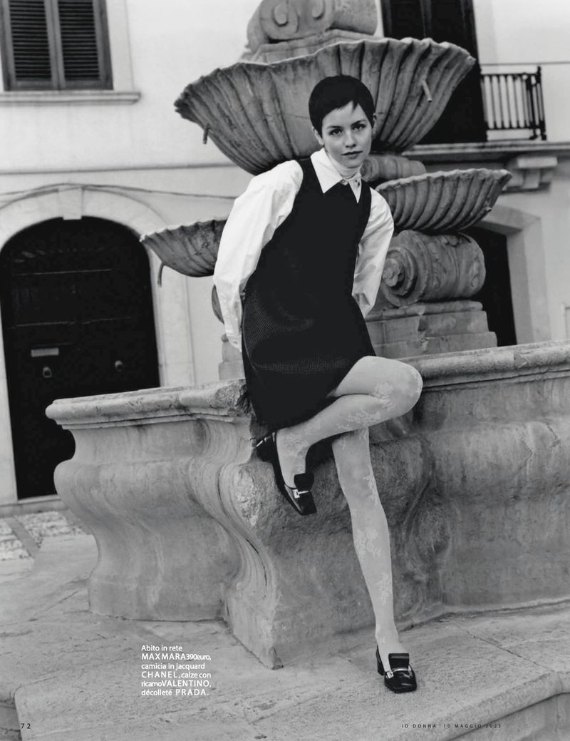 Monochrome fashion shot of a young woman in a dress and blouse sitting on a stone fountain outdoors with a building in the background.