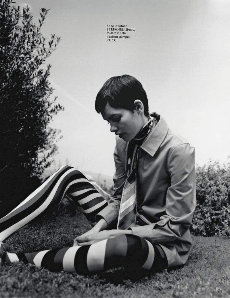 Monochrome fashion shot of a woman in striped leggings and a coat, sitting on grass in a natural outdoor setting with trees and sky.