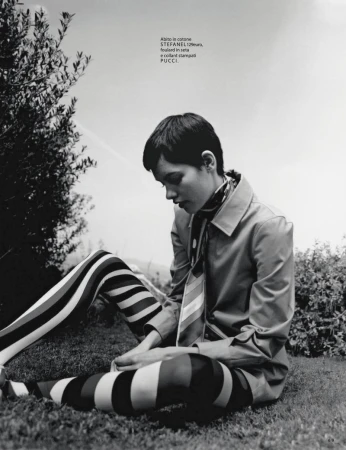 Monochrome fashion shot of a woman in striped leggings and a coat, sitting on grass in a natural outdoor setting with trees and sky.