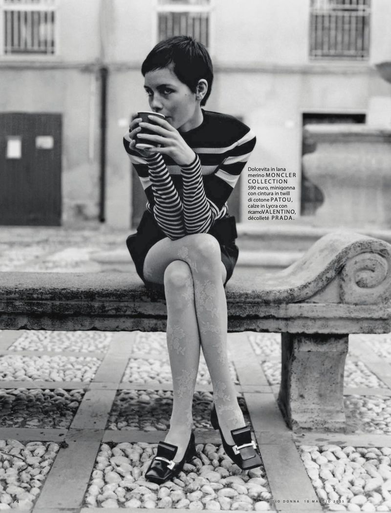 A woman with short hair, wearing a striped shirt, is sitting on a stone bench and drinking from a cup in an outdoor setting.
