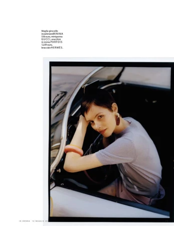 A woman with short hair sits in the driver's seat of a vintage car, wearing a lavender top and bracelet, looking at the camera.