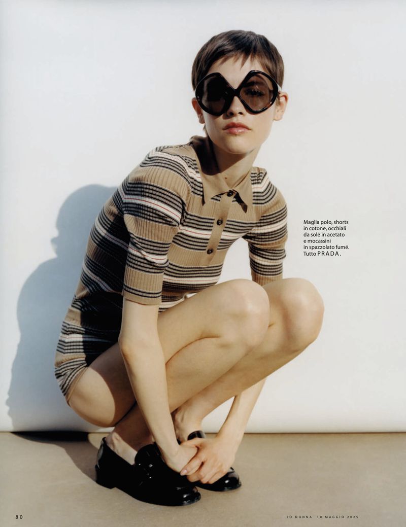 Young woman in striped polo dress and black loafers, squatting in a photo studio, styled with oversized dark sunglasses, fashion editorial.