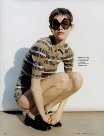 Young woman in striped polo dress and black loafers, squatting in a photo studio, styled with oversized dark sunglasses, fashion editorial.