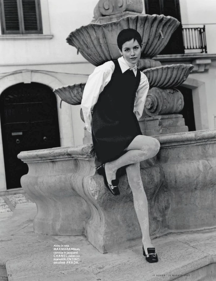 Model with short dark hair wearing a dark pinafore dress and white shirt seated on a stone fountain for iO Donna.