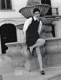 Model with short dark hair wearing a dark pinafore dress and white shirt seated on a stone fountain for iO Donna.