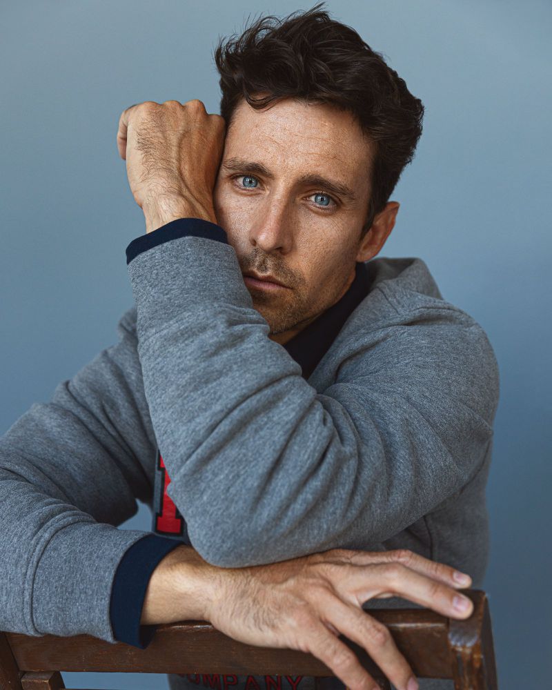 Adult man with blue eyes sits with hands resting on a wooden chair wearing gray hooded sweatshirt in portrait against blue background.