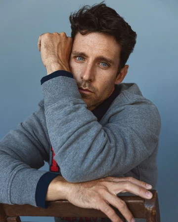 Adult man with blue eyes sits with hands resting on a wooden chair wearing gray hooded sweatshirt in portrait against blue background.