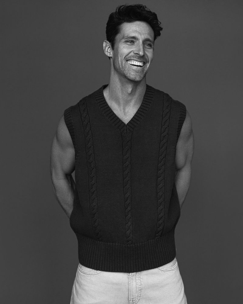Black and white portrait of a smiling adult male wearing a knitted vest and light-wash jeans against a solid grey backdrop.