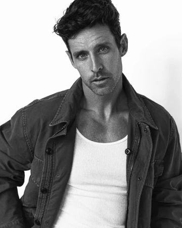 Monochrome portrait of a brunette adult male with olive skin wearing a white ribbed tank top and jacket against a white backdrop.