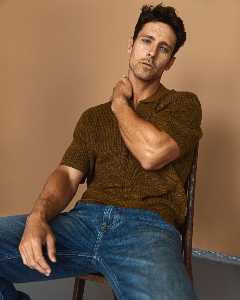 Portrait of a handsome man in a polo shirt and jeans sitting on a chair against a neutral tan background in a studio setting.