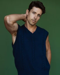 Man wearing a navy blue cable-knit sweater vest posing against a solid dark green studio background