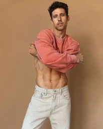 Tyler Lough pulling off a coral red sweatshirt against a warm beige background for The Rakish Gent editorial project by Blake Ballard