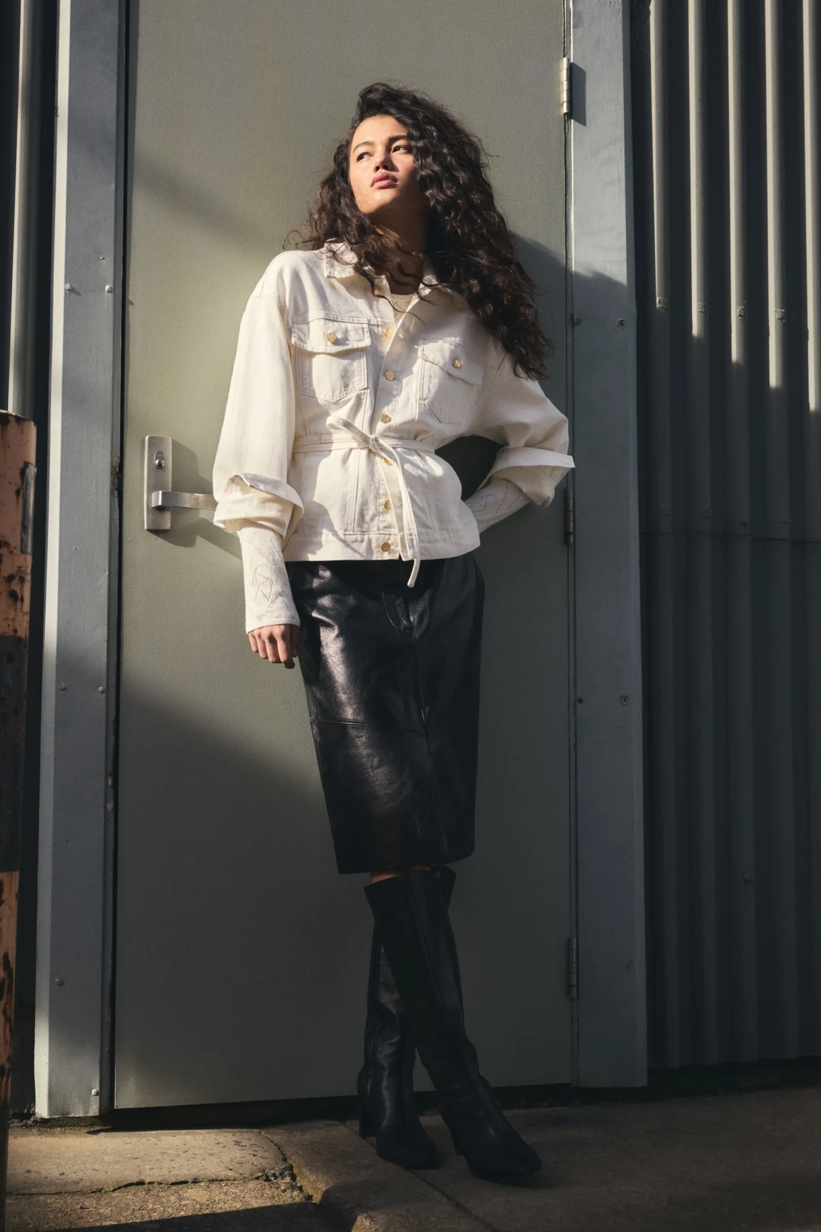 A stylish young woman with curly hair, wearing a white denim jacket, black skirt, and boots, poses against a neutral door outside.