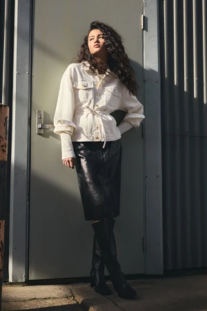 A stylish young woman with curly hair, wearing a white denim jacket, black skirt, and boots, poses against a neutral door outside.