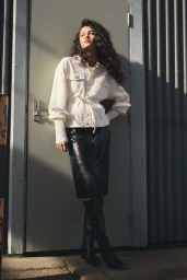 Model with long dark curly hair wearing a white belted jacket and black leather skirt standing outdoors