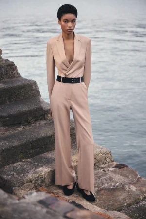 Adult female model in a beige pant suit with a belt stands on stone steps near the sea in an outdoor fashion editorial photoshoot.