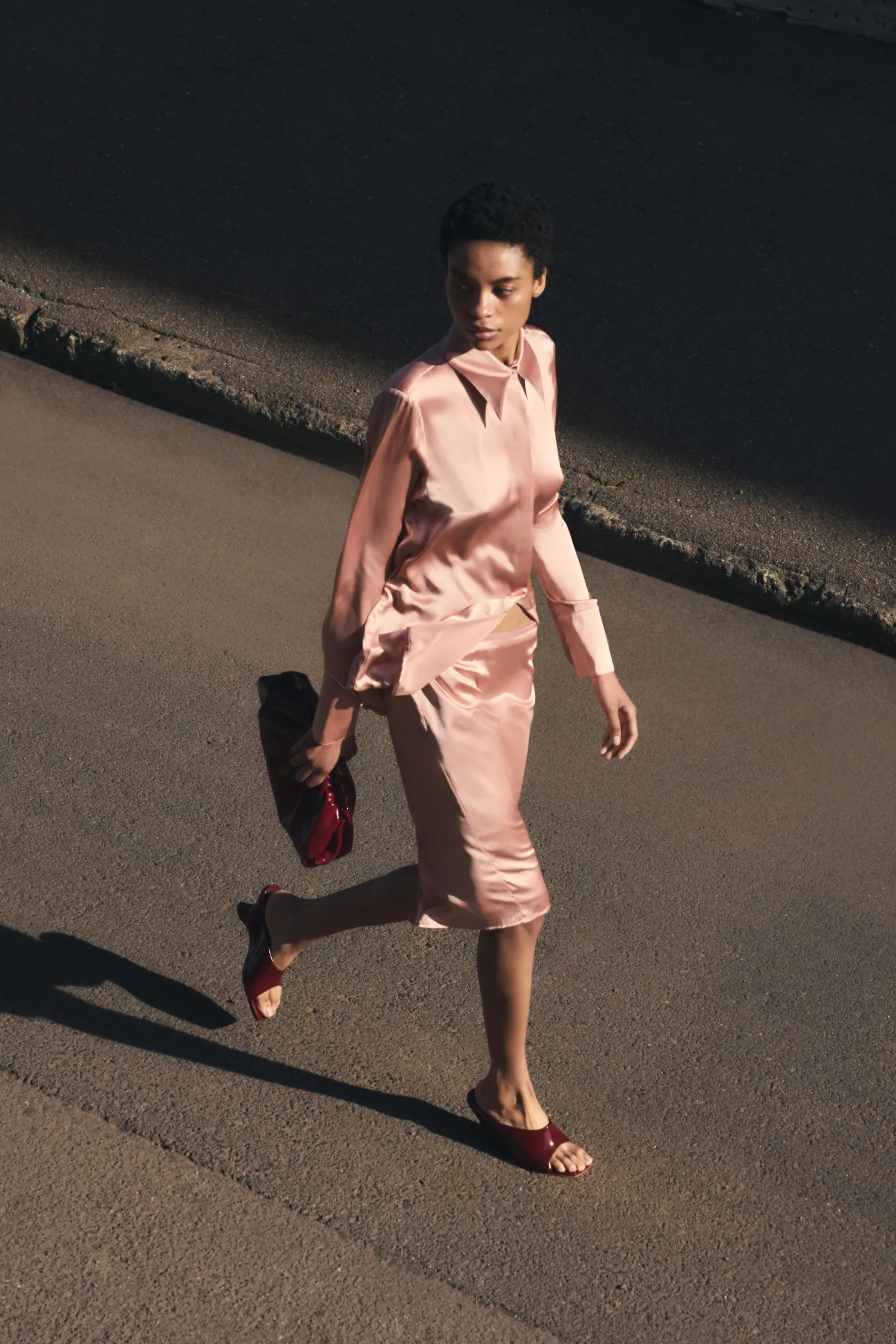 Fashionable adult woman in pink satin dress, matching purse, and slide sandals strides confidently along sunlit urban street.